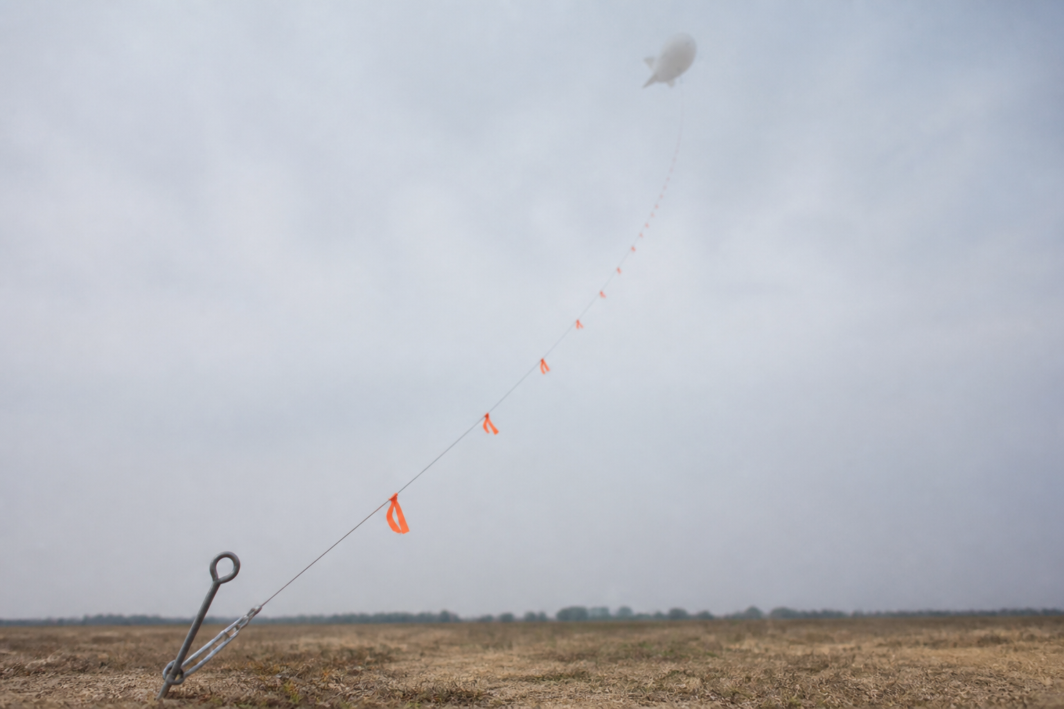 A tether line rising from a ground anchor into a hazy sky with bright orange fabric pennants tied along it at regular intervals, a small aerostat visible as a pale shape high above, illustrating the marking of a mooring line